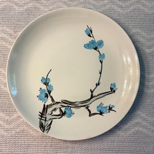 May include: A round, off-white ceramic plate featuring a hand-painted design of a dark brown tree branch with bright blue blossoms. The branch extends from the lower left side, curving upward and across the center of the smooth, circular surface.