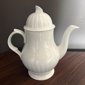 May include: A white ceramic teapot with a lid, handle, and spout. The teapot has a rounded body with vertical ridges and sits on a small pedestal base. The lid has a knob on top. The teapot is on a wooden surface.