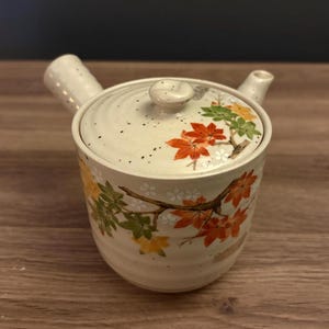 May include: A ceramic kyusu teapot with a side handle and a lid, featuring a cream-colored base with speckled details and a painted design of orange, red, and green maple leaves on both the body and the lid, including a small spout.
