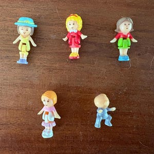 Könnte beinhalten: Fünf kleine Kunststofffiguren. Eine trägt einen blauen Hut und ein gelbes Kleid. Eine hat gelbes Haar und ein rotes Outfit. Eine hat braunes Haar, ein rotes Oberteil und eine grüne Hose. Eine hat orangefarbenes Haar, ein rosa Oberteil und einen Rock. Eine ist eine blaue Babyfigur.