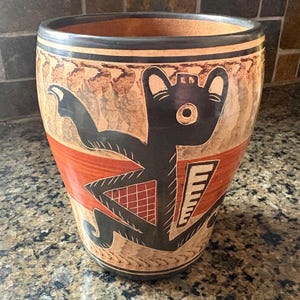 May include: A ceramic vase with a wide, cylindrical shape. The vase features a tan background with a black and red design of a stylized figure. The figure has a large head and outstretched arms. The vase has a dark brown rim.