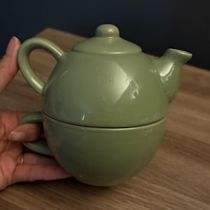 May include: A sage green ceramic tea set consisting of a teapot with a lid and a matching cup stacked underneath. The teapot features a curved handle and a short spout, while the cup has a small handle, both with a smooth, glossy finish.
