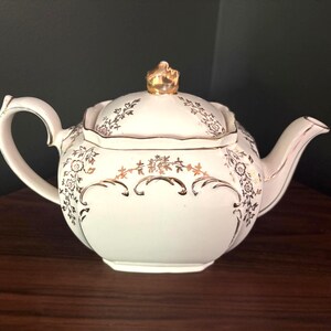May include: A white ceramic teapot featuring a gold floral pattern and gold trim along the edges. The teapot has a curved handle, a spout, and a lid topped with a gold-colored finial. The body of the teapot has a scalloped, octagonal shape.