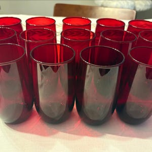 May include: A collection of fourteen ruby red drinking glasses. The glasses are cylindrical with a slightly rounded base. The glasses are arranged on a white surface. The glasses are made of a transparent material.