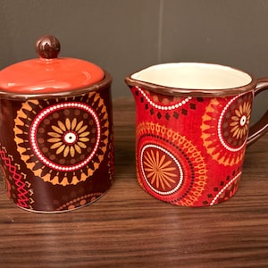 May include: A ceramic sugar bowl with a dark brown base and a bright orange lid, paired with a matching creamer pitcher featuring a vibrant red and orange circular pattern. Both pieces display a consistent geometric design with brown rims.