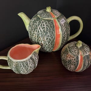 May include: A ceramic tea set featuring a teapot, a creamer, and a sugar bowl, all designed to resemble a melon. The pieces have a textured dark green exterior with a white cross-hatched pattern and a smooth orange vertical stripe.