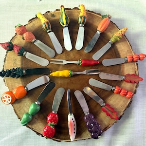 May include: A collection of fifteen metal spreaders and appetizer forks featuring colorful, three-dimensional handles shaped like various fruits, vegetables, and leaves, including strawberries, grapes, oranges, corn, and pumpkins.