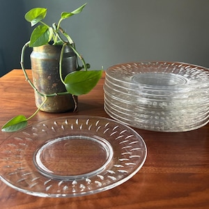 May include: A stack of clear glass plates with a decorative pattern, next to a single plate and a small, aged metal container holding a green plant. The plates have a textured design around the edges.