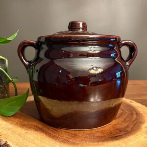 May include: A glossy, dark brown ceramic bean pot with a lid and two side handles. The pot sits on a wooden slice, with a small plant in a dark container to the left. The pot's surface reflects light.