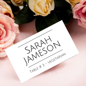 以下が含まれることがあります： 黒い文字で「SARAH JAMESON」と書かれた白い席札。その下に「TABLE # 3 - VEGETARIAN」と書かれています。カードはピンクの皿の上にあり、ピンクと黄色のバラに囲まれています。