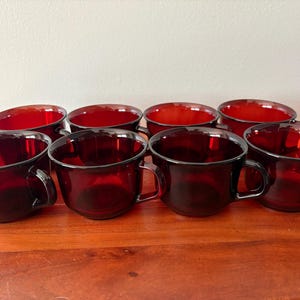 May include: Eight ruby red glass cups with handles are arranged on a wooden surface. The cups have a rounded shape with a dark rim and a handle on the side. The cups are empty and appear to be clean.