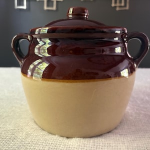 May include: A two-handled ceramic bean pot with a lid, featuring a glossy dark brown glaze on the upper portion and a matte tan finish on the lower body. The pot has a rounded shape with two side handles and a small knob on the center of the lid.