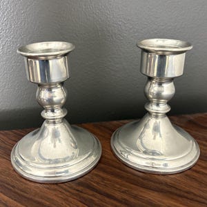 May include: Two silver-colored metal candlesticks. Each candlestick has a round base, a bulbous center, and a cylindrical top to hold a candle. The candlesticks are displayed on a dark wooden surface.