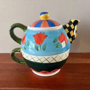 May include: A colorful ceramic teapot with a matching teacup base. The teapot features a light blue body with red tulip designs, a green handle, and a lid with orange and blue stripes. The spout has a black and yellow checkered pattern.