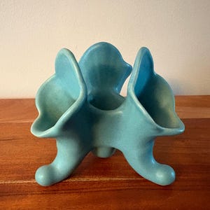 May include: A turquoise ceramic decorative object with three curved sections and three rounded feet. The object has a smooth, matte finish and a unique, abstract design. It is likely a vase or a decorative piece.