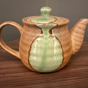 May include: A ceramic teapot with a textured, light brown body and a smooth, pale green glaze accent running down the center. It features a matching lid with a small knob, a curved handle on the left, and a ridged spout on the right side.