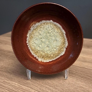 May include: A round ceramic bowl featuring a deep reddish-brown outer rim and a textured, crystalline center in shades of pale yellow and white. The bowl is displayed on a clear plastic stand, showcasing its circular shape and contrasting glaze.