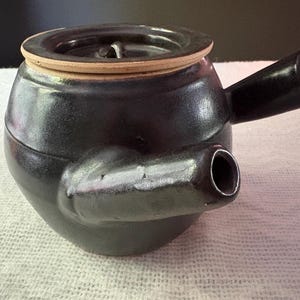 May include: A dark charcoal-colored ceramic teapot with a side-mounted handle and a short, protruding spout. The teapot features a circular lid with a light beige rim and a small central knob, showcasing a smooth, glossy finish throughout the body.