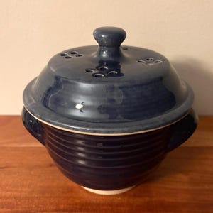 May include: A dark blue ceramic bowl with a matching lid featuring a knob handle and four sets of small ventilation holes. The bowl has horizontal ribbed detailing along the sides and two small integrated handles on opposite sides of the base.