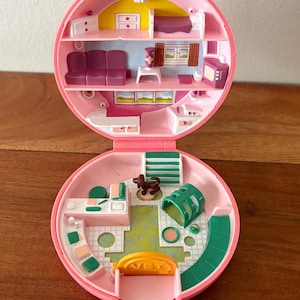 May include: Pink vintage toy playset, open to reveal a miniature house with multiple rooms. The interior features a bedroom, living room, and a vet clinic. Includes a brown dog figurine and green and yellow accents.