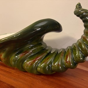 May include: A ceramic cornucopia centerpiece featuring a glossy, dark forest green glaze with subtle brown and amber highlights. The horn-shaped vessel has a wide, open mouth and a spiraled, tapered tail, showcasing a smooth, reflective finish.
