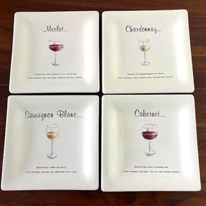 May include: A set of four square white ceramic appetizer plates. Each plate features a unique wine glass illustration and text identifying the wine variety: Merlot, Chardonnay, Sauvignon Blanc, and Cabernet, along with descriptive tasting notes.