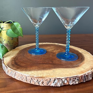 May include: Two clear martini glasses with blue beaded stems and bases sit on a round wooden serving board. The glasses have a classic triangular bowl shape. A potted plant with green leaves is in the background.