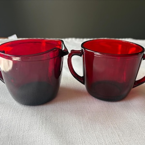 May include: Two ruby red glass serving pieces. One is a creamer with a spout and handle. The other is a sugar bowl with two handles. Both pieces are made of a dark red glass and sit on a white surface.