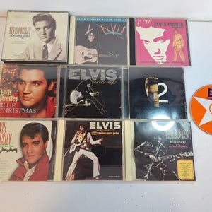 May include: A collection of Elvis Presley CDs, featuring album covers with images of Elvis, including "Amazing Grace," "Blue Christmas," and "Viva Las Vegas." The CDs are in their original jewel cases, with a single orange CD on the right.