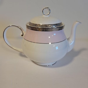 May include: A white porcelain teapot featuring a soft pink horizontal band around the upper body, accented by a decorative silver metallic trim on the lid and rim. The teapot includes a curved handle, a spout, and a matching lid with a knob.