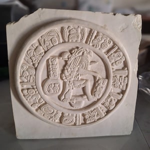 May include: A square off-white stone plaque featuring a circular relief carving. The center depicts a seated figure in profile, surrounded by a ring of intricate glyphs and symbols. The surface has a matte, textured finish with visible carved details.
