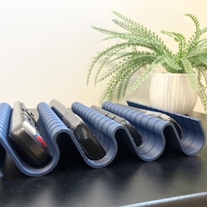 May include: A blue, wavy remote control organizer holding several remotes. The organizer is made of a flexible material and has a series of curved slots to hold the remotes. A decorative plant in a white pot is in the background.