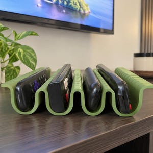 Peut inclure: Un organisateur de télécommandes vert olive au design ondulé à plusieurs fentes. Le support présente quatre canaux verticaux distincts, chacun occupé par une télécommande électronique noire, offrant une solution de rangement structurée.