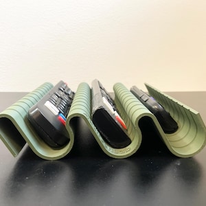 Könnte beinhalten: Ein salbeigrüner Fernbedienungsorganizer mit wellenförmigem Design, der vier schwarze Fernbedienungen hält. Der Organizer hat eine gerippte Textur und besteht aus einem flexiblen Material. Die Fernbedienungen haben verschiedene Tasten und sind unterschiedlich groß.