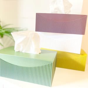 Peut inclure: Trois boîtes à mouchoirs rectangulaires présentant une texture nervurée verticale sur les côtés et une découpe incurvée lisse sur le devant. Les boîtes sont de couleur vert sauge, blanc éclatant et mauve profond, avec une base jaune visible.