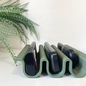 Puede incluir: Un soporte organizador de color verde salvia con forma de onda que cuenta con múltiples ranuras diseñadas para sostener mandos a distancia electrónicos en posición vertical. El soporte tiene un acabado mate y una estructura curva.