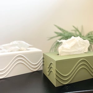 以下が含まれることがあります： 前面に波模様のデザインが施された、白とオリーブグリーンの長方形のティッシュボックスカバーが2つ。各ボックスには白いティッシュの束が入っています。ボックスは、背景に植物がある暗い表面にあります。