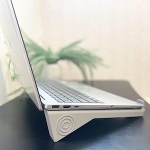 May include: A silver laptop is supported by a grey stand with a circular pattern on its side. The laptop is open, displaying the keyboard. The stand rests on a black surface. The background is blurred.