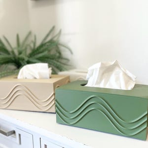 Peut inclure: Deux boîtes à mouchoirs rectangulaires, l'une vert sauge et l'autre beige clair, présentant toutes deux un motif en relief composé de lignes ondulées sur la face avant, avec des mouchoirs blancs dépassant de l'ouverture supérieure.