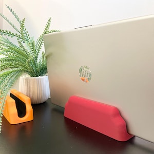 May include: A silver HP laptop is held by a red stand. An orange stand and a potted plant with green leaves are also visible. The laptop has the HP logo on the back.
