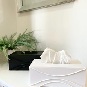 May include: Two tissue boxes, one black and one white, sit side-by-side on a white surface. The white box has a decorative wave design. A bundle of white tissues is pulled from the white box. A green plant in a pot is in the background.