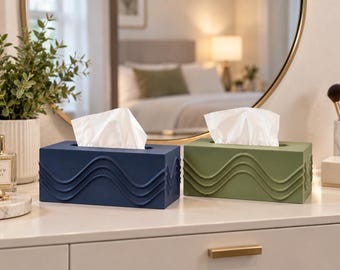 May include: Two rectangular tissue box covers, one in navy blue and one in olive green, each featuring a raised, horizontal wavy line pattern across the front and sides. Both covers are open at the top, with white tissues protruding from the centre.