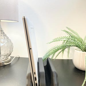 May include: A silver laptop sits in a black laptop stand. The stand is made of a dark material and has a U-shaped design. The laptop is angled, showing its side profile with ports. A lamp and a potted plant are in the background.