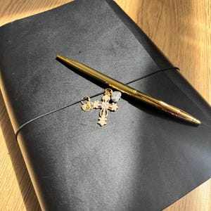 Op de afbeelding: Een zwart notitieboek met lederen kaft, vastgezet met een zwarte elastische band. Een goudkleurige balpen ligt diagonaal over de kaft. Aan de elastische band hangt een decoratieve goudkleurige kruishanger met kleine doorzichtige steentjes.