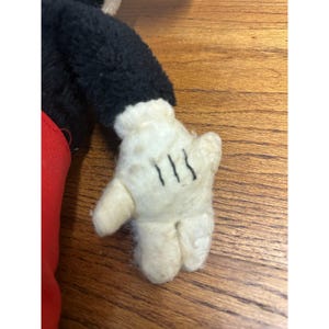 May include: Close-up of a vintage Mickey Mouse plush toy's white glove with three black lines on the palm. The glove is attached to a black sleeve and red fabric. The plush is on a wooden surface.