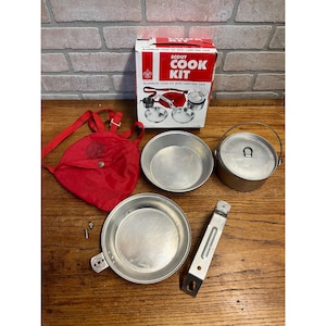 May include: Vintage Scout Cook Kit with carrying case. The kit includes a red carrying bag, a box with the text "SCOUT COOK KIT", two aluminum pans, a pot with a handle, and a metal handle. The box reads "ALUMINUM COOK KIT WITH CARRYING CASE".