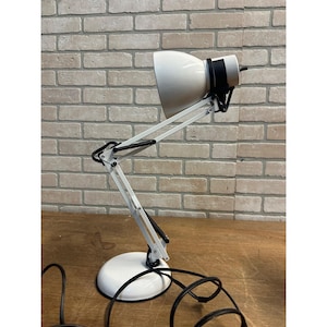 May include: White desk lamp with a black and white articulated arm and a round base. The lamp has a conical shade and a black power cord. The lamp is on a wooden surface and in front of a brick wall.