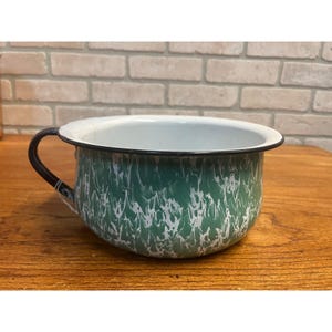 May include: A vintage enamelware bowl with a green and white swirled pattern. The bowl has a black rim and handle. The interior is white. The bowl is sitting on a wooden surface.