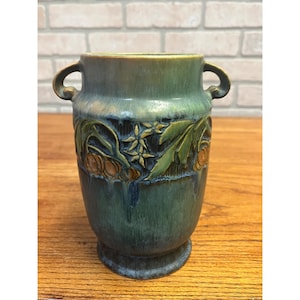 May include: A ceramic vase featuring a mottled green and blue glaze with a relief pattern of leaves and round fruit. The vessel has two curved handles on the sides, a wide opening, and a flared base, showcasing a textured finish throughout.
