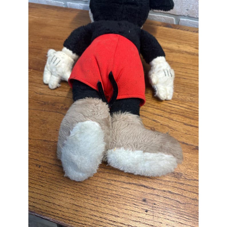 May include: A vintage Mickey Mouse soft toy, seen from behind. It has a black body, red shorts, white gloves, and large, two-tone brown and white shoes. The toy is placed on a wooden surface.